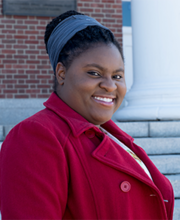Daniela Belice standing on the steps of Boyden Hall smiling at the camera with her black hair pulled up in a bun and wearing a red coat with lapel and buttons