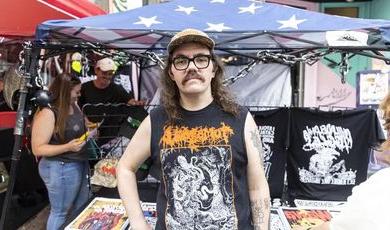 A man with glasses wearing a hat stands in front of a tent selling tshirts 