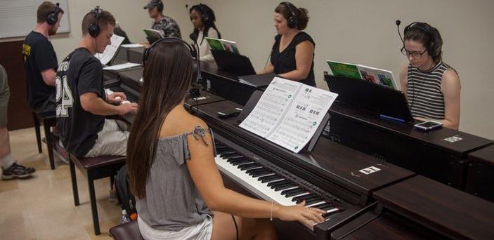 八架钢琴背靠背地放在教室中央，每架钢琴上都有一名学生戴着耳机弹奏.