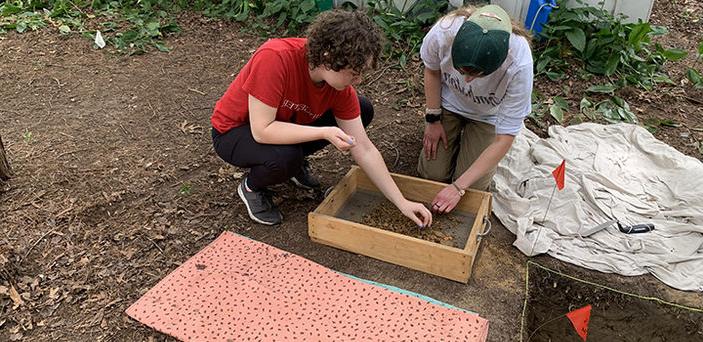 2 BSU archeology students kneel on a plot of dirt picking through items in a sieve taken from a hole they dug in the ground