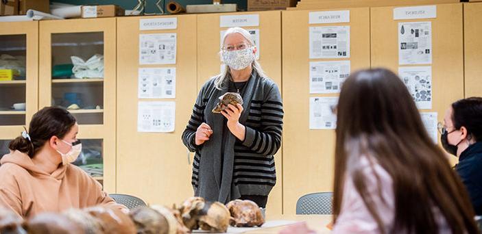 BSU Professor Ellen Ingmanson holds a skull while she teaches at the head of a table with a row of skulls on it that students sit around listening and taking notes